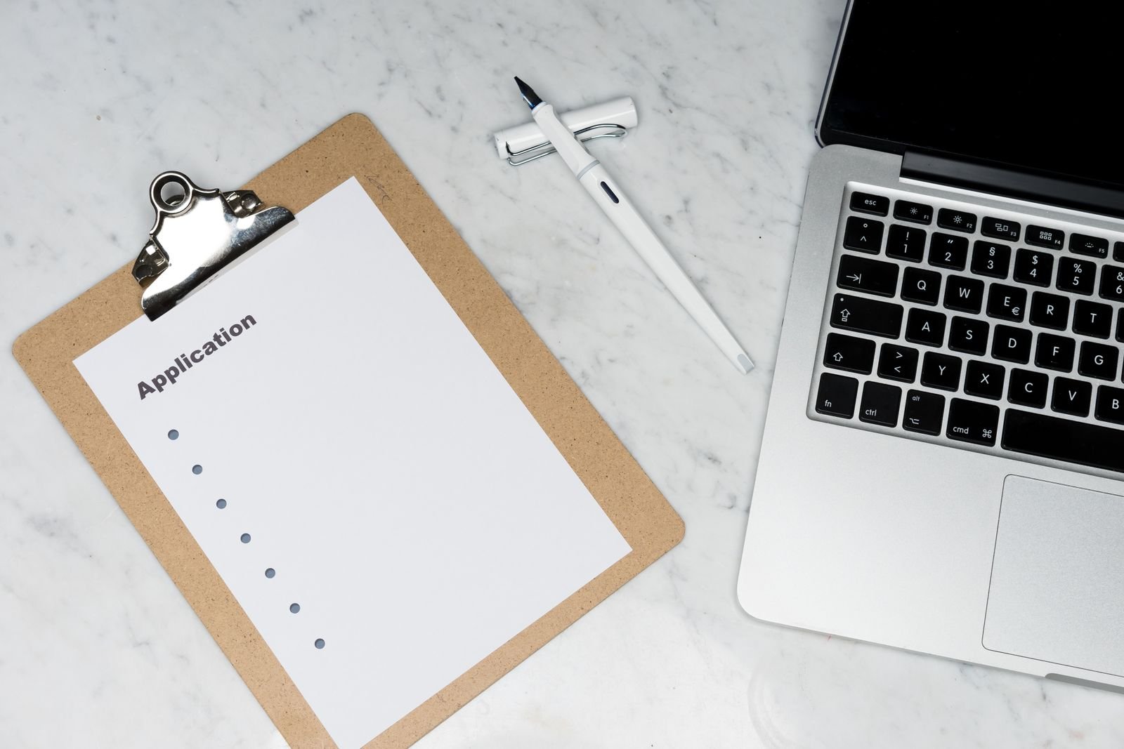 A blank application form on a clipboard beside a pen and an open laptop on a desk