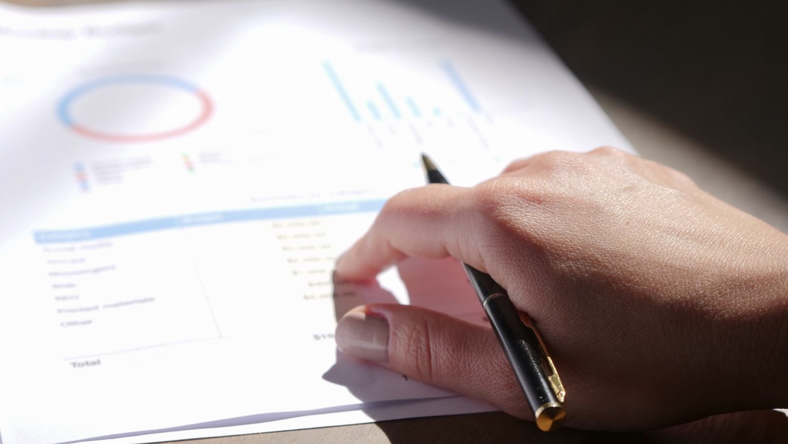 A hand holding a pen over printed financial charts and figures on a desk